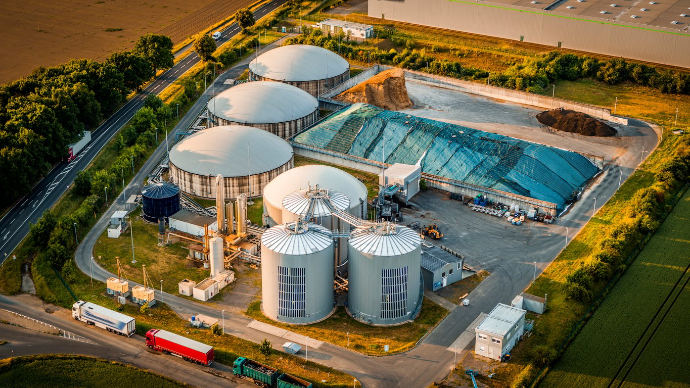 Aerial view of a modern biogas facility in golden light, highlighting its large silos, covered feedstock storage, and circular fermentation tanks. Located near agricultural fields , the photo emphasizes the intersection of sustainable energy and rural infrastructure